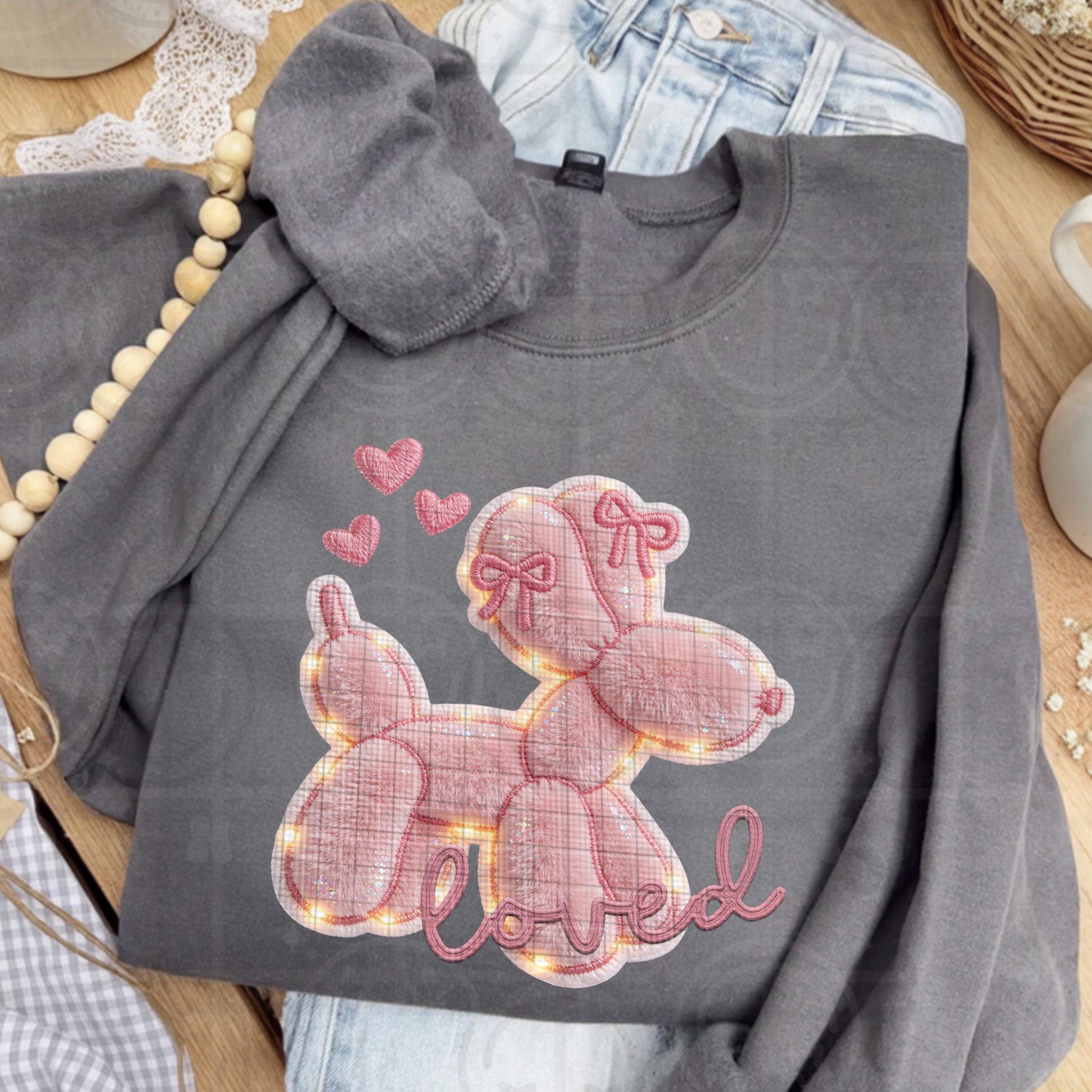 The shirt design shows an adorable pink balloon dog with rounded segments, accented by small bows and floating hearts above its head. The artwork mimics layered applique fabric with faux embroidery outlines, subtle faux fur texture, and soft glowing faux lights around the edges, finished with the word “loved” in stitched-style script beneath the dog.