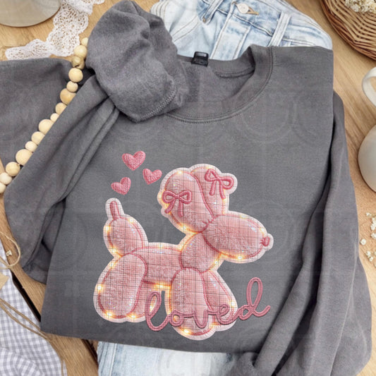 The shirt design shows an adorable pink balloon dog with rounded segments, accented by small bows and floating hearts above its head. The artwork mimics layered applique fabric with faux embroidery outlines, subtle faux fur texture, and soft glowing faux lights around the edges, finished with the word “loved” in stitched-style script beneath the dog.
