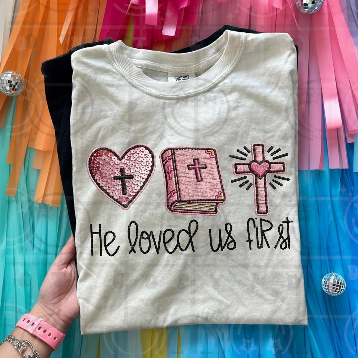 Faith based faux embroidery design reading He Loved Us First with stitched style heart bible and cross icons in pink tones
